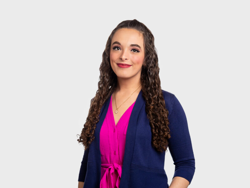 Jenny Eyler, a woman with dark brown hair wearing a warm fuchsia dress with a dark blue cardigan.