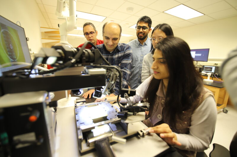 Several STEM students are gathered around equipment in a lab, with a young woman working on a project in the foreground and a professor looking over her shoulder.