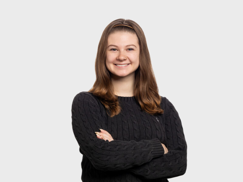 Abigail Ozog, a young woman with brown hair, a black sweater, and her arms folded across her chest. She is smiling at the camera.