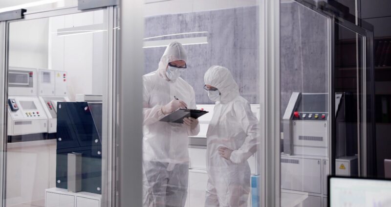 Fully gowned personnel inside a cleanroom reviewing data on a clipboard among specialized laboratory equipment.