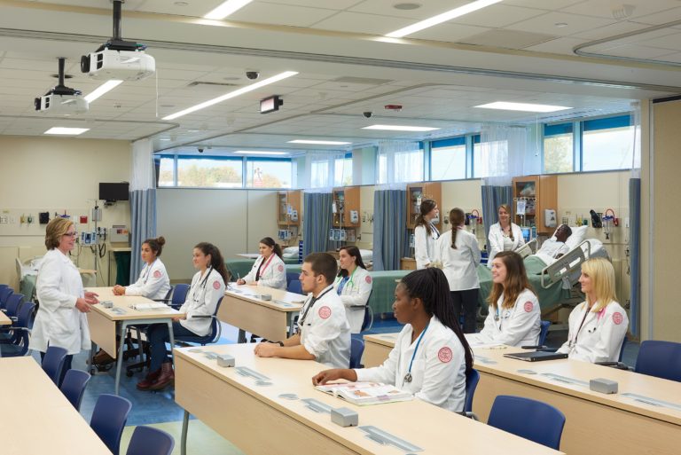 A group of health education students sit in a lecture hall, attentively listening to their instructor. All are wearing lab coats.