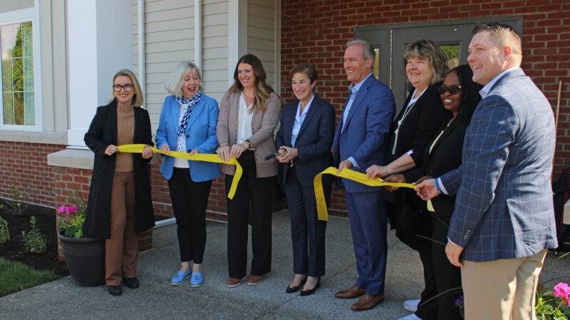 A group of people stand in front of a brick building at a ribbon-cutting.