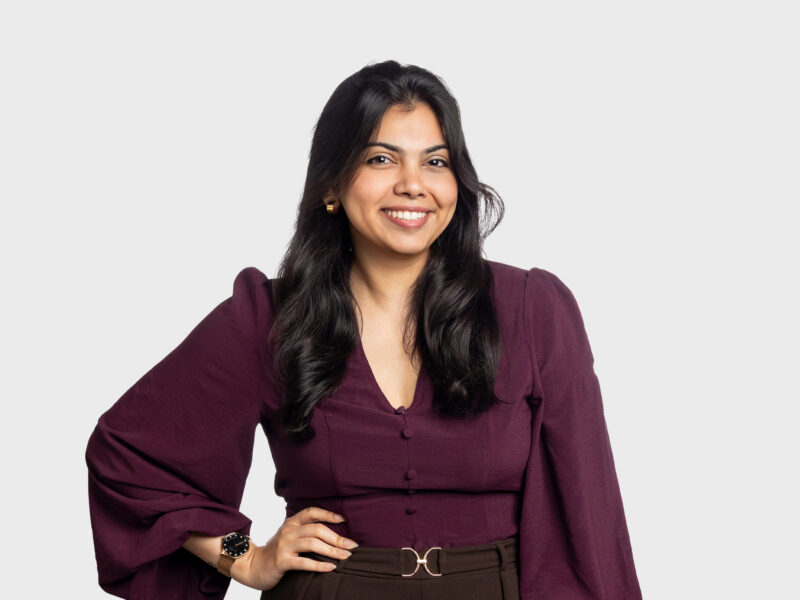 A photo of Devika Kharkar, a young woman with a burgundy blouse and dark hair. She stands with one hand on her hip, looking at the camera.
