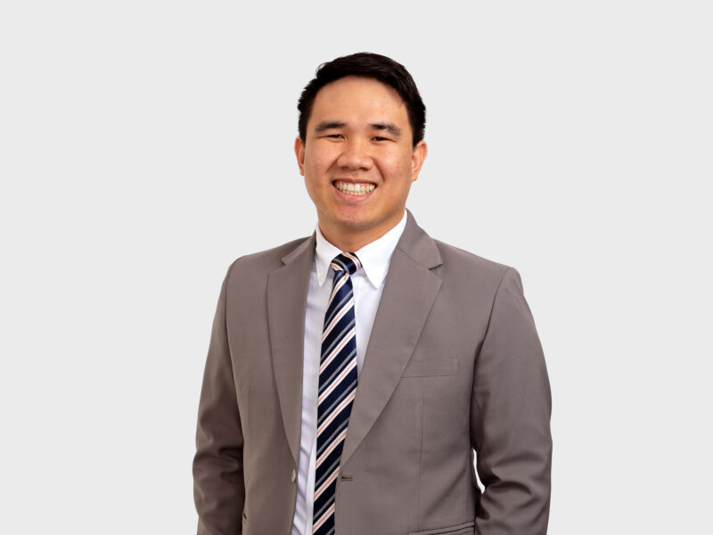 A photo of J. Lawrence Manalo, who is wearing a gray suit with a blue and white striped tie. He is smiling at the camera, looking straight ahead. The background is plain white.