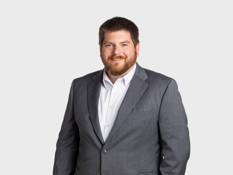 A photo of Benjamin Deuel, who is wearing a gray suit with a white dress shirt. He is smiling at the camera, looking straight ahead. The background is plain white.