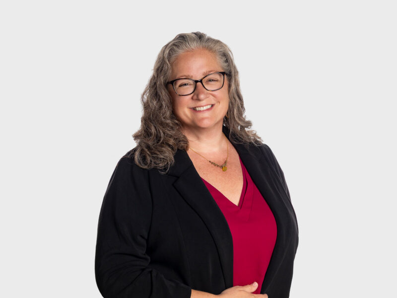 A photo of Meredith Gifford-Morath, who is wearing a red blouse with a black cardigan. She is smiling at the camera, looking straight ahead. The background is plain white.